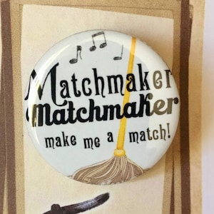 May include: A white button with black text that reads "Matchmaker Matchmaker make me a match!" with a yellow broom and musical notes. The button is in a frame with the text "Fiddler on the Roof" below it.