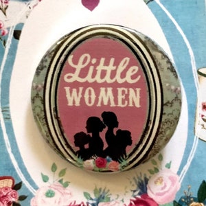 May include: A round button with a pink background and white text that reads "Little Women". The button is surrounded by a floral design and a white banner with the text "Little Women" in pink.