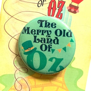 Peut inclure: Un bouton vert avec le texte "The Merry Old Land of Oz" et un petit chapeau vert avec une bande dorée. Le bouton est sur un fond jaune et orange avec une montgolfière et le texte "We're off to see the Wizard... The Wonderful Wizard of Oz".
