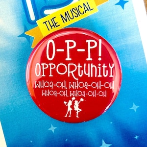 May include: A red button with the text "O-P-P! OPPORTUNITY" and the words "WHOA-OH WHOA-OH-OH WHOA-OH WHOA-OH-OH" in smaller text below. The button is in front of a card with the number "13" and the text "THE MUSICAL" on it.