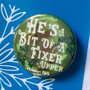 May include: A green button with the text "He's a Bit of a Fixer Upper" and a green ivy design. The button is on a blue background with a white floral design.