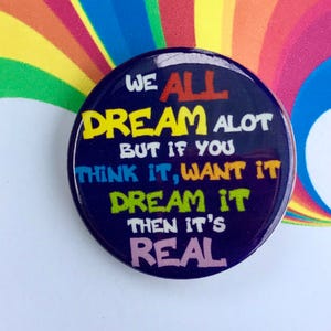 Puede incluir: Un botón azul con el texto "WE ALL DREAM ALOT BUT IF YOU THINK IT, WANT IT DREAM IT THEN IT'S REAL" en letras de colores. El botón está en una tarjeta blanca con un fondo de arcoíris y el texto "JOSEPH AND THE AMAZING TECHNICOLOR DREAMCOAT".
