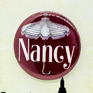 May include: A burgundy button featuring a white umbrella and the word "Nancy" in white text. The button is displayed on a card with the text "OLIVER!" in black and a silhouette of a city skyline.