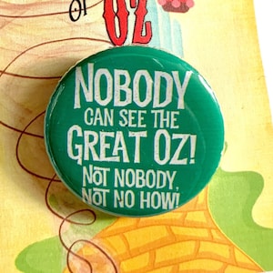 Peut inclure: Un bouton vert avec le texte "Nobody can see the Great Oz! Not nobody, not no how!" sur un fond jaune avec une montgolfière et une route de briques jaunes.