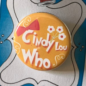 Op de afbeelding: Een gele knop met de tekst "Cindy Lou Who" in rood-witte letters. Er is een rode strik en witte bloemen op de knop. De knop bevindt zich op een witte kaart met een blauwe rand en een zwart-witte illustratie van een hoed.