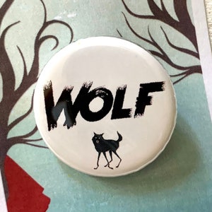 May include: White button with black text that says "WOLF" and a small black wolf silhouette. The button is on a card with the text "INTO THE WOODS THE MUSICAL".