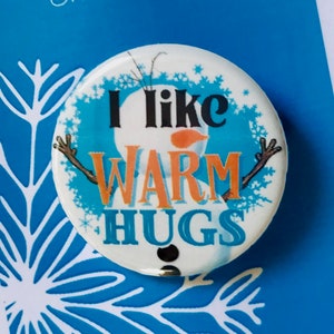 May include: A white button with a blue background featuring a cartoon snowman with the text "I like warm hugs". The button is on a blue card with the text "Frozen The Musical".