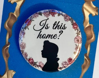 May include: A white button with a floral border and a silhouette of Belle from Beauty and the Beast. The button says "Is this home?" and is on a blue background with a gold frame.