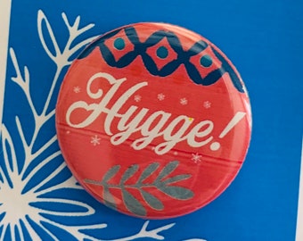 May include: A red button with a white snowflake design and the text "Hygge!" in white. The button is on a blue card with a white snowflake design and the text "Frozen The Musical" in white.