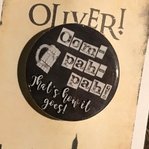 May include: A black button with a white image of a beer mug and the text "oom-pah-pah! That's how it goes!"  The button is on a card with the text "Oliver!" and a black silhouette of the London skyline.