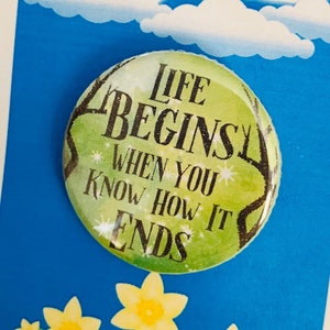 Puede incluir: Un botón verde con el texto "Life Begins When You Know How It Ends" sobre un fondo azul con nubes blancas y el texto "Big Fish". El botón está rodeado de narcisos amarillos.