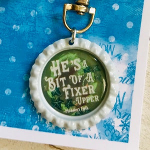 May include: A white bottle cap keychain with a green and white design. The bottle cap has the text "He's a bit of a fixer upper" on it. The keychain is attached to a silver metal key ring.