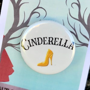 May include: A white button with the text "Cinderella" in black and a yellow glass slipper. The button is on a card with the text "Into the Woods The Musical" in black.