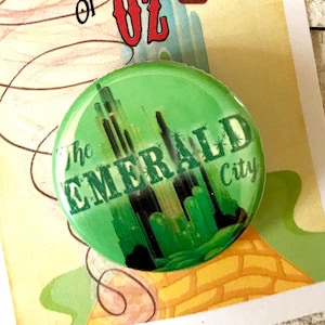 Peut inclure: Un bouton vert avec le texte "The Emerald City" et une image stylisée de la Cité d'Émeraude du Magicien d'Oz. Le bouton est sur une carte avec le texte "We're off to see the Wizard... The Wonderful Wizard of Oz" et une montgolfière.
