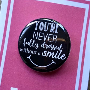 Puede incluir: Un botón negro con texto blanco que dice "You're never fully dressed without a smile". El botón está en una tarjeta rosa con el texto "Annie the musical" en blanco.