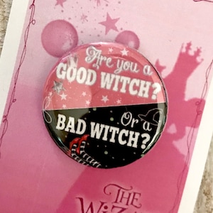 Peut inclure: Un bouton rose et noir avec le texte "Êtes-vous une bonne sorcière ? Ou une mauvaise sorcière ?" et les mots "Le Magicien d'Oz" sur un fond rose.