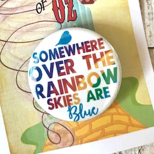 Peut inclure: Une impression avec des images du Magicien d'Oz, avec un bouton épinglé dessus. L'impression présente une montgolfière et le texte "We're off to see the Wizard". Le bouton dit "Somewhere Over the Rainbow Skies are Blue."