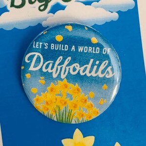 Puede incluir: Un botón azul y blanco con un diseño de narcisos amarillos. El botón dice "Let's build a world of Daffodils". El botón está sobre un fondo azul con nubes blancas y las palabras "Big Fish" en letras verdes.