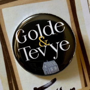 Peut inclure: Un bouton noir avec du texte blanc qui dit "Golde & Tevye" et une petite illustration d'une maison. Le bouton est sur une carte avec le texte "Fiddler on the Roof" en écriture noire.