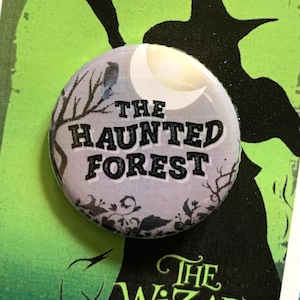 Peut inclure: Un bouton rond avec un motif noir et blanc. Le bouton représente une lune, des arbres et le texte "The Haunted Forest". Le bouton est sur une carte avec une silhouette de la Méchante Sorcière de l'Ouest du Magicien d'Oz.