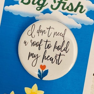 Puede incluir: Un botón blanco con el texto "I don't need a roof to hold my heart" en escritura negra. El botón está sobre un fondo azul con una nube blanca y el texto "Big Fish" en verde. Hay narcisos amarillos en la parte inferior de la imagen.