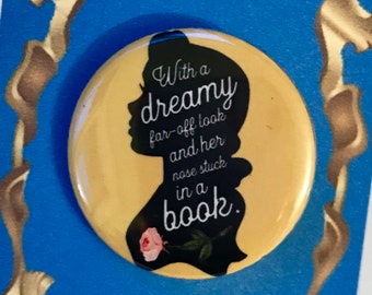 May include: A yellow button with a black silhouette of Belle from Beauty and the Beast with the text "With a dreamy far-off look and her nose stuck in a book." The button is on a blue background with a gold frame and the text "Beauty and the Beast".