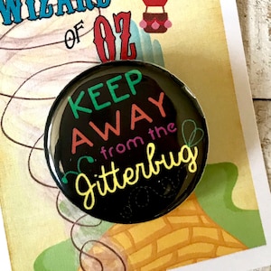 Peut inclure: Un bouton noir avec le texte "Keep Away from the Jitterbug" en lettres colorées. Le bouton est devant une carte avec une montgolfière et le texte "We're off to see the Wizard... The Wonderful Wizard of Oz."