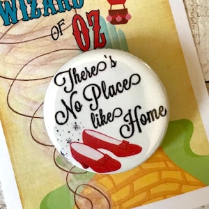 Peut inclure: Un bouton blanc avec le texte "There's No Places like Home" en lettres noires avec des pantoufles rouges en rubis sur un fond de route de briques jaunes. Le bouton est attaché à une carte avec une montgolfière et le texte "We're off to see the Wizard... The Wonderful Wizard of Oz."
