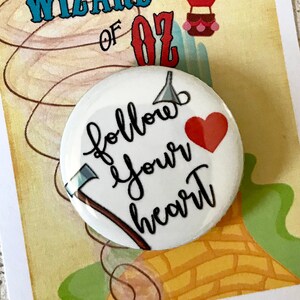 K&ouml;nnte beinhalten: Ein wei&szlig;er Knopf mit dem Text "follow your heart" in schwarzer Schrift. Ein rotes Herz und eine silberne Axt befinden sich ebenfalls auf dem Knopf. Der Knopf befindet sich auf einer Karte mit einem Cartoon-Bild eines Hei&szlig;luftballons und eines Tornados. Die Karte sagt "We're off to see the Wizard... The Wonderful Wizard of Oz."