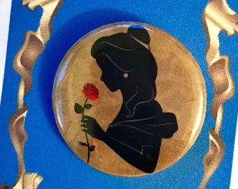 May include: A round button featuring a silhouette of Belle from Beauty and the Beast, holding a red rose. The button is on a blue background with a gold frame and the text "Beauty and the Beast".