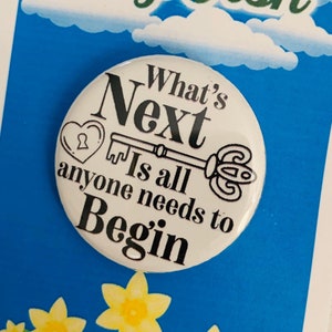 Könnte beinhalten: Ein weißer Knopf mit schwarzem Text, der "What's Next Is all anyone needs to Begin" lautet. Der Knopf hat eine Schlüssel- und Herzgrafik. Der Knopf befindet sich auf einem blauen Hintergrund mit weißen Wolken und gelben Blumen.
