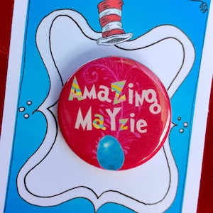 May include: A red button with the text "Amazing Mayzie" in white and yellow lettering. The button has a blue and white design in the background. The button is on a white card with a blue background and a black and white illustration of a hat.