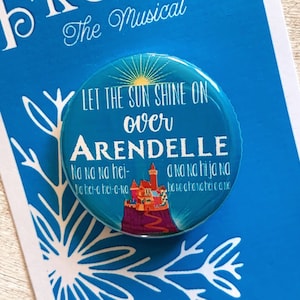 May include: A blue and white button with a picture of a castle and the text "Let the sun shine on over Arendelle" with lyrics from the song "Let it Go" from the musical Frozen.