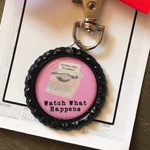 Puede incluir: Un llavero de tapón de botella rosa y negro con una imagen de máquina de escribir y el texto "Watch What Happens". El llavero está atado con una cinta roja.