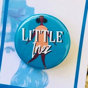 May include: A blue button with a white outline and the text "LITTLE Inez" in white. The button features a cartoon illustration of a woman with brown skin and a pink dress. The button is on a white card with the text "hairspray" in colorful letters.