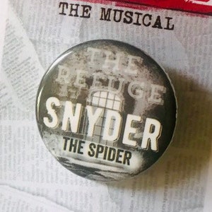Puede incluir: Un botón blanco y negro con el texto "The Refuge Snyder The Spider" en él. El botón está en una tarjeta blanca con el texto "Newsies The Musical" en ella.