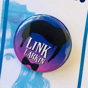 May include: A blue, pink, and black button with a silhouette of a person's head and the text "LINK LARKIN" in white. The button is on a white card with the word "hairspray" in rainbow colors.