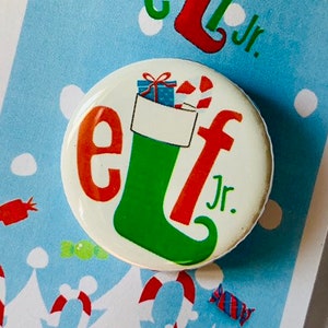 May include: A white button with a green and red elf boot with a gift and candy cane inside. The text "elf Jr." is on the button. The button is on a white card with a blue background and a winter scene.
