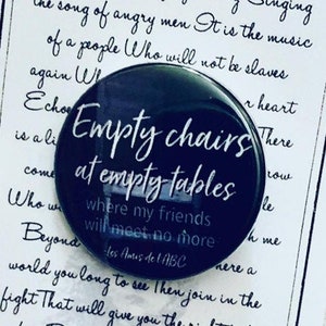 May include: A black button with white text that reads "Empty chairs at empty tables where my friends will meet no more - Les Amis de l'ABC". The button is on a white card with text from the musical Les Misérables.