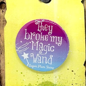 May include: A round button with a pink, purple, and blue ombre design. The button features white text that reads "They broke my Magic Wand" and "Sugar Plum Fairy". The button is attached to a yellow background with a quote from Shrek that reads "What makes us special, makes us strong."