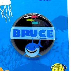 Op de afbeelding: Een Finding Nemo Jr. knoop met een blauwe haai met witte tanden en het woord "BRUCE" in blauwe letters op een blauwe achtergrond.
