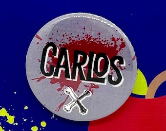 May include: A grey button with a red splatter design and the word "CARLOS" in black lettering. The button also features a white crossbones design. The button is on a blue background with yellow splatter designs and a red apple.