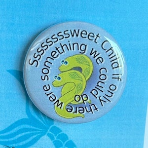 Peut inclure: Un bouton bleu clair avec un fond blanc et une image de dessin animé de deux anguilles. Le bouton porte le texte "Ssssssweet Child if only we could do there were something". Le bouton est sur une carte bleu clair avec le texte "Disney's The Little Mermaid" en blanc.
