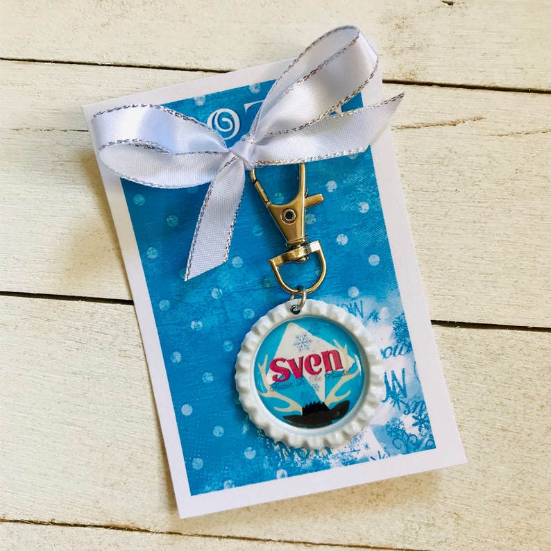 May include: A white bottle cap keychain with a blue and pink design featuring the word "Sven" and a reindeer silhouette. The keychain is attached to a silver key ring and a white ribbon.