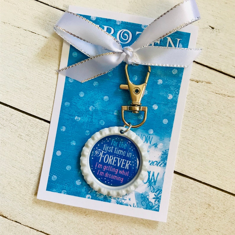 K&ouml;nnte beinhalten: Ein wei&szlig;er Flaschenverschluss-Schl&uuml;sselanh&auml;nger mit blauem Hintergrund und dem Text "For the first time in FOREVER I'm getting what I'm dreaming". Der Schl&uuml;sselanh&auml;nger ist an einem silbernen Schl&uuml;sselring befestigt und wird auf einem blauen Hintergrund mit wei&szlig;en Punkten gezeigt.