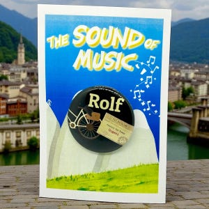 May include: A postcard featuring the title "The Sound of Music" with a black button that says "Rolf" and a bicycle graphic. A small telegram is attached to the button. The background shows a mountain scene.