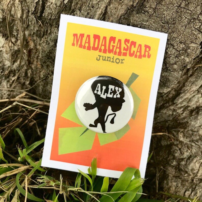 May include: A white button featuring a black silhouette of Alex the lion from the Madagascar films. The button reads "Alex" in white lettering. The button is displayed against a yellow background with the text "Madagascar Junior" in black.