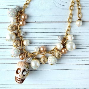 Puede incluir: Collar de cadena dorada con una variedad de elementos decorativos. El collar presenta un colgante de calavera, cuentas de perlas y esferas con incrustaciones de cristal. El collar tiene una etiqueta que dice "Pierre Dumas, NYC, Original Design."