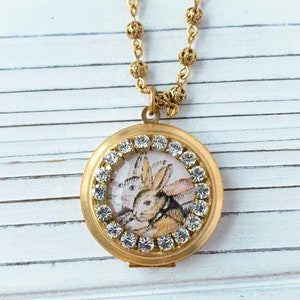 Puede incluir: Collar de medallón dorado con una ilustración de un conejo blanco rodeado de brillantes transparentes.