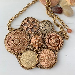 May include: A gold chain necklace with a large pendant made of beige and brown ceramic buttons. The buttons are intricately carved with floral and geometric designs. The necklace is made by Lenora Dame and is made in the USA.